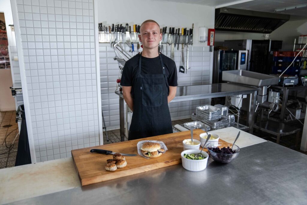 Ung slagter står bag træbræt med burger, frikadeller og salater.