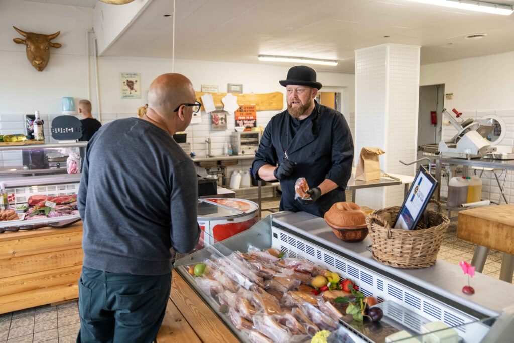 Slagter i sort uniform taler med kunde ved køledisken.